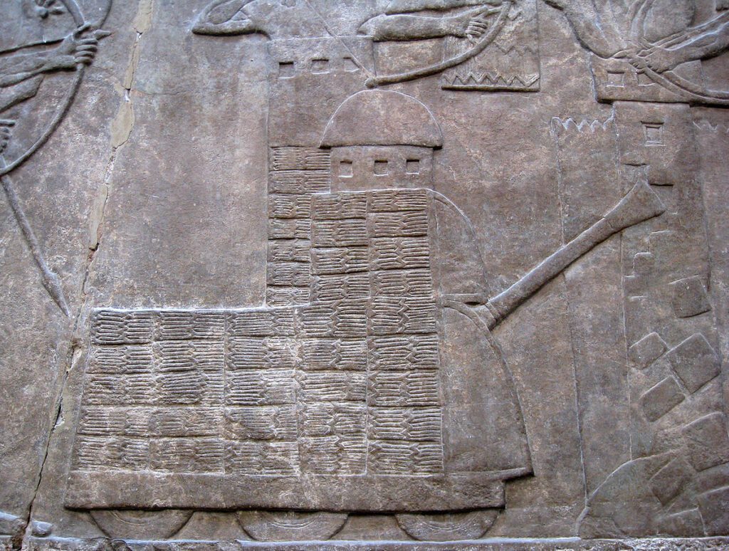 Assyrian relief showing a wheeled battering ram with protective superstructure attacking city walls.